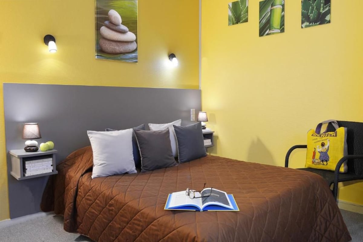 A bedroom with a brown bedspread, two pillows, a bedside lamp, a nightstand with books and a lamp, and a wall-mounted light fixture.