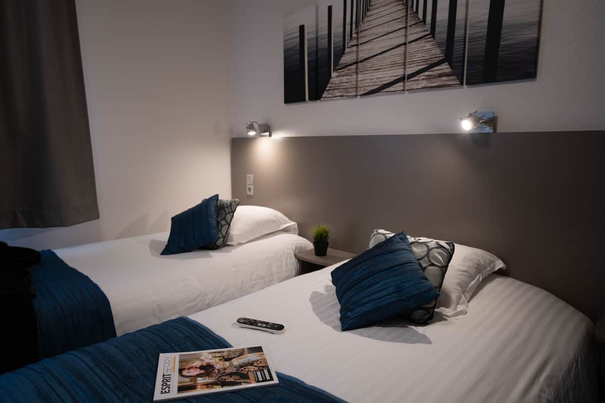A hotel room with two beds, each with a pillow and a remote control. A magazine is on the bed.