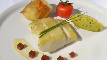 Un plato con un trozo de pescado blanco, un tomate, una rebanada de pan frito y una porción pequeña de patata.