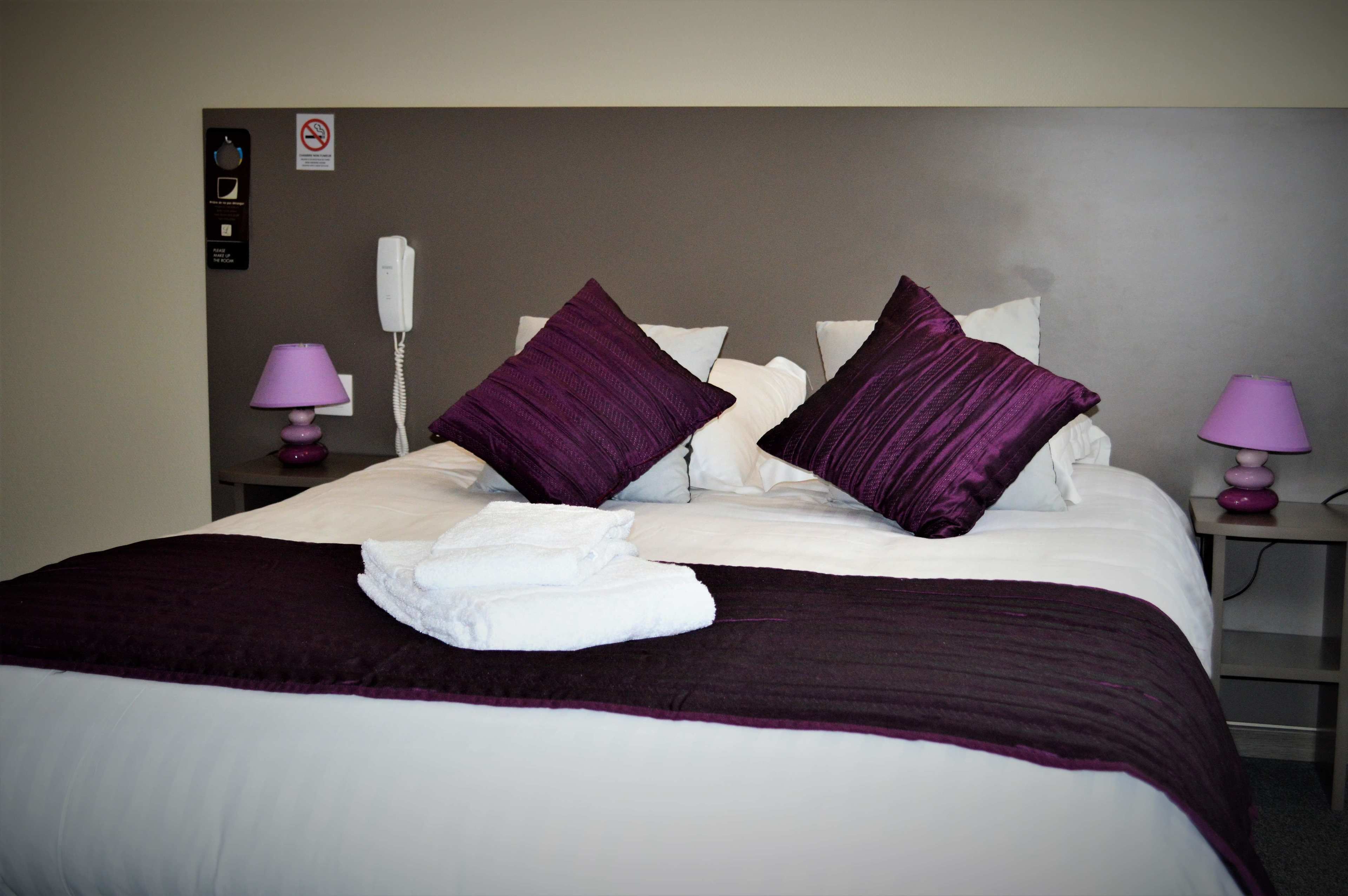 A neatly made bed with a purple blanket and pillows, a white bedside table with a lamp, and a phone on the wall.