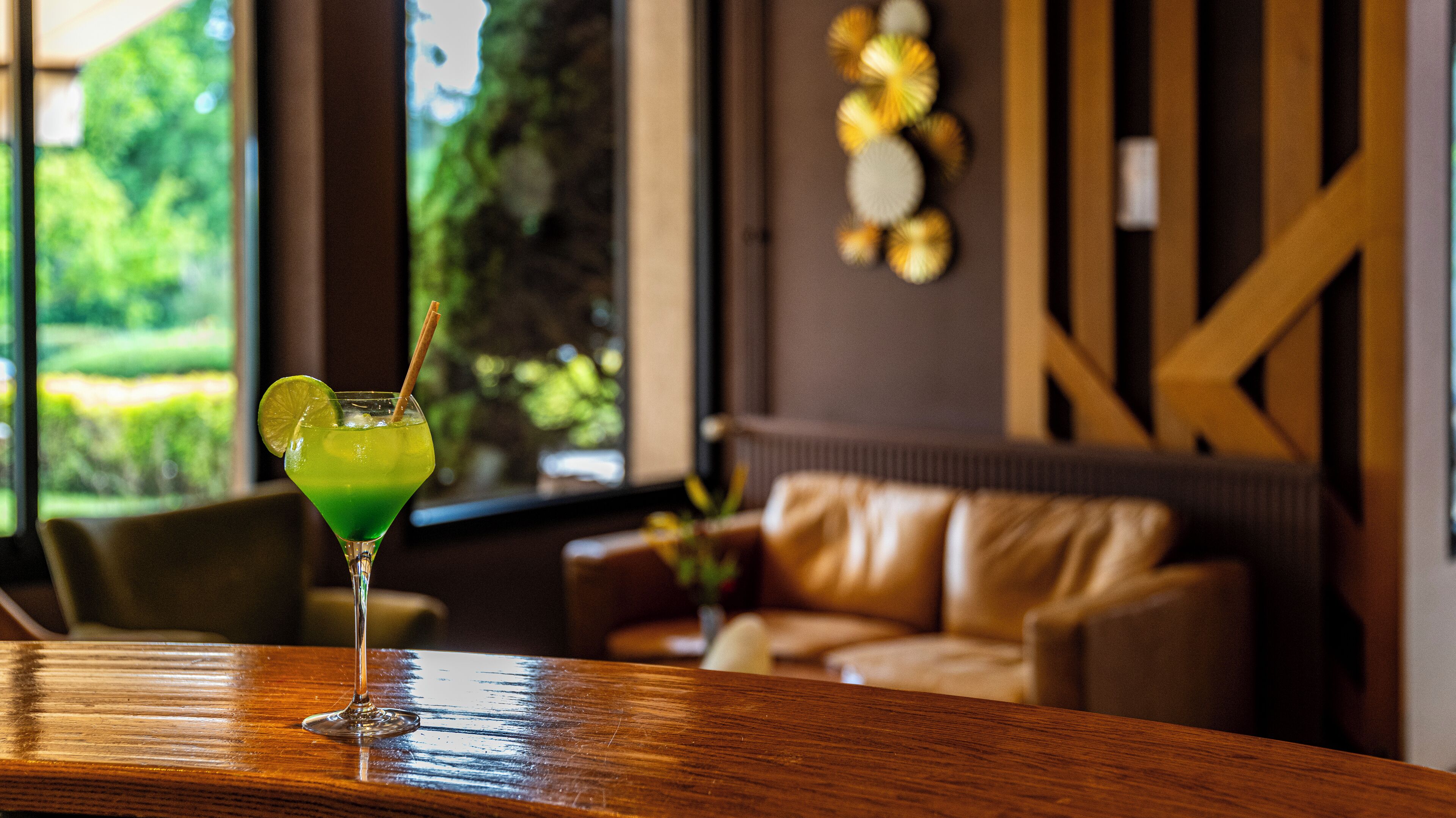 Een bar met een groene cocktail op een houten oppervlak, een leren bank en uitzicht op groen door grote ramen.