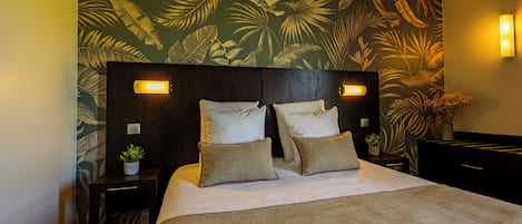 Habitación de hotel con una cama grande, dos mesitas de noche, un papel tapiz con hojas tropicales y dos lámparas de pared.