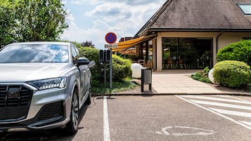 Un coche aparcado en un estacionamiento, con un cartel de ‘prohibido estacionar’, la entrada de un edificio y una zona de estar al aire libre techada.