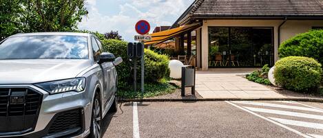 Un coche aparcado en un estacionamiento, con un cartel de ‘prohibido estacionar’, la entrada de un edificio y una zona de estar al aire libre techada.