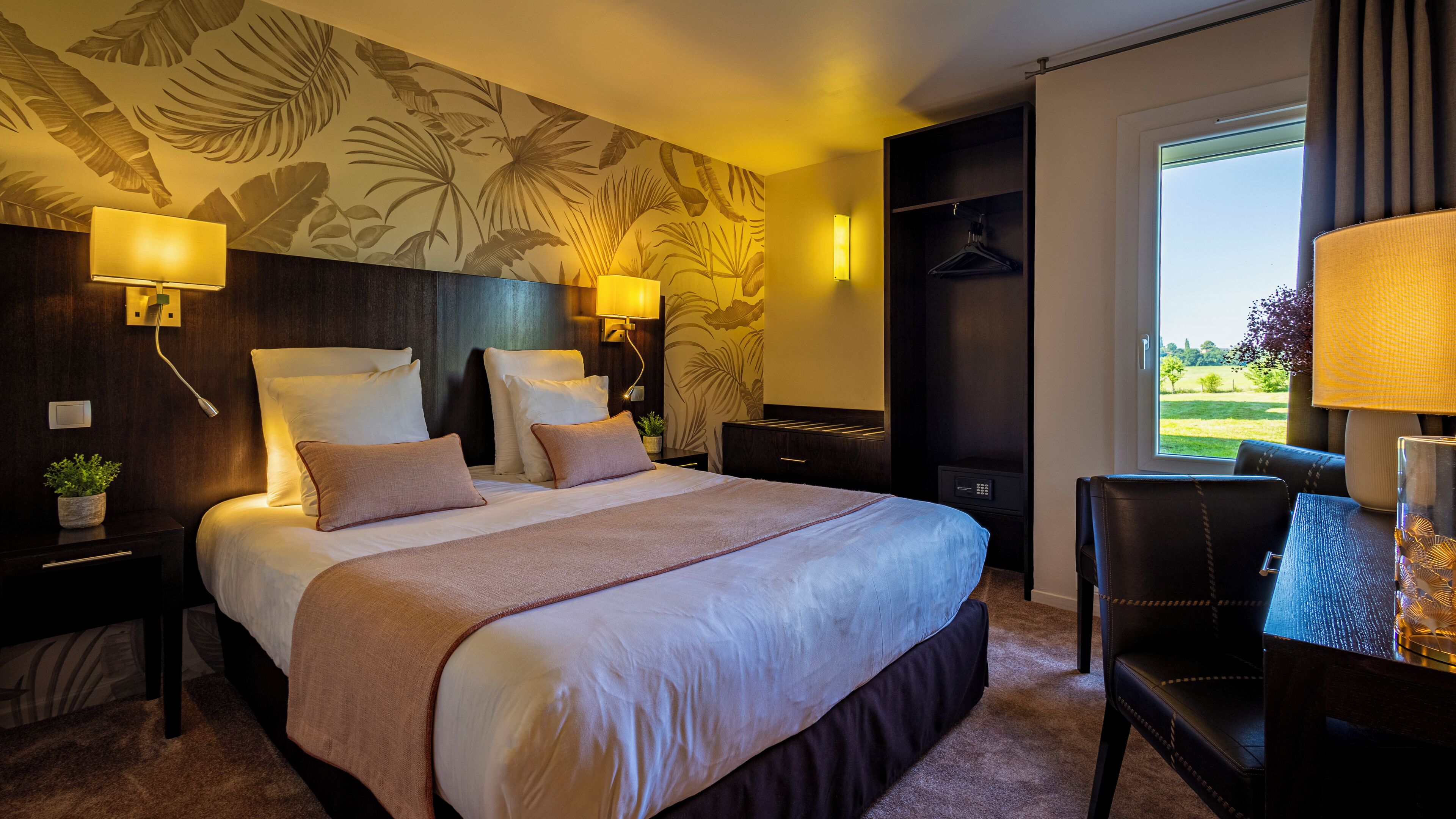 A hotel room with a large bed, two bedside tables with lamps, a dark headboard, a large window with a view of greenery, and a seating area with a chair and table.