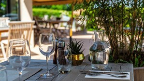 Una mesa dispuesta con copas de vino, una botella de agua y una jarrón, con una planta y asientos exteriores al fondo.
