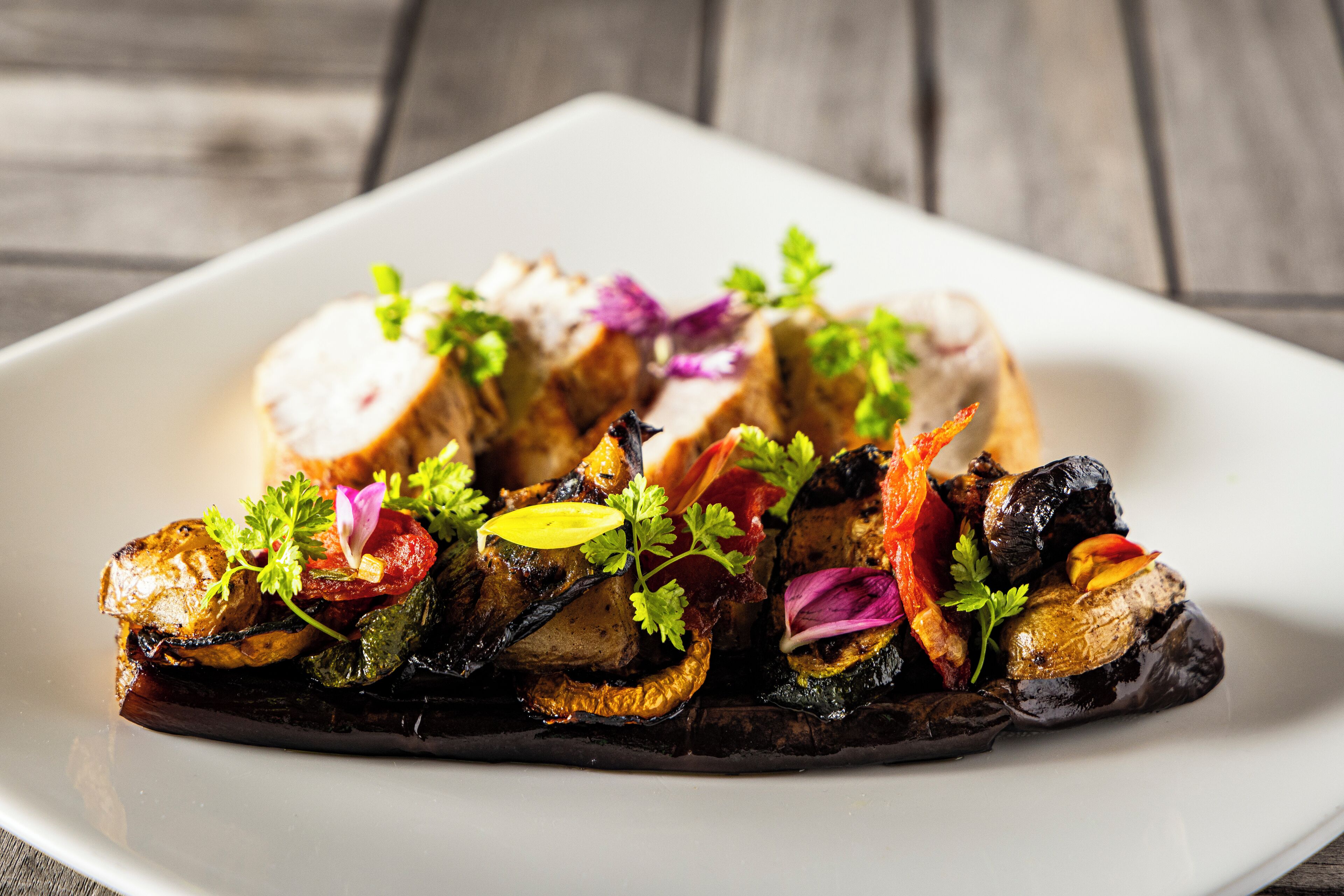 A white plate with a roasted chicken, eggplant, and vegetables garnished with herbs.