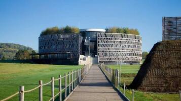 Un bâtiment moderne doté d’une façade géométrique originale, un chemin en bois menant à l’entrée et, en arrière-plan, un champ verdoyant.