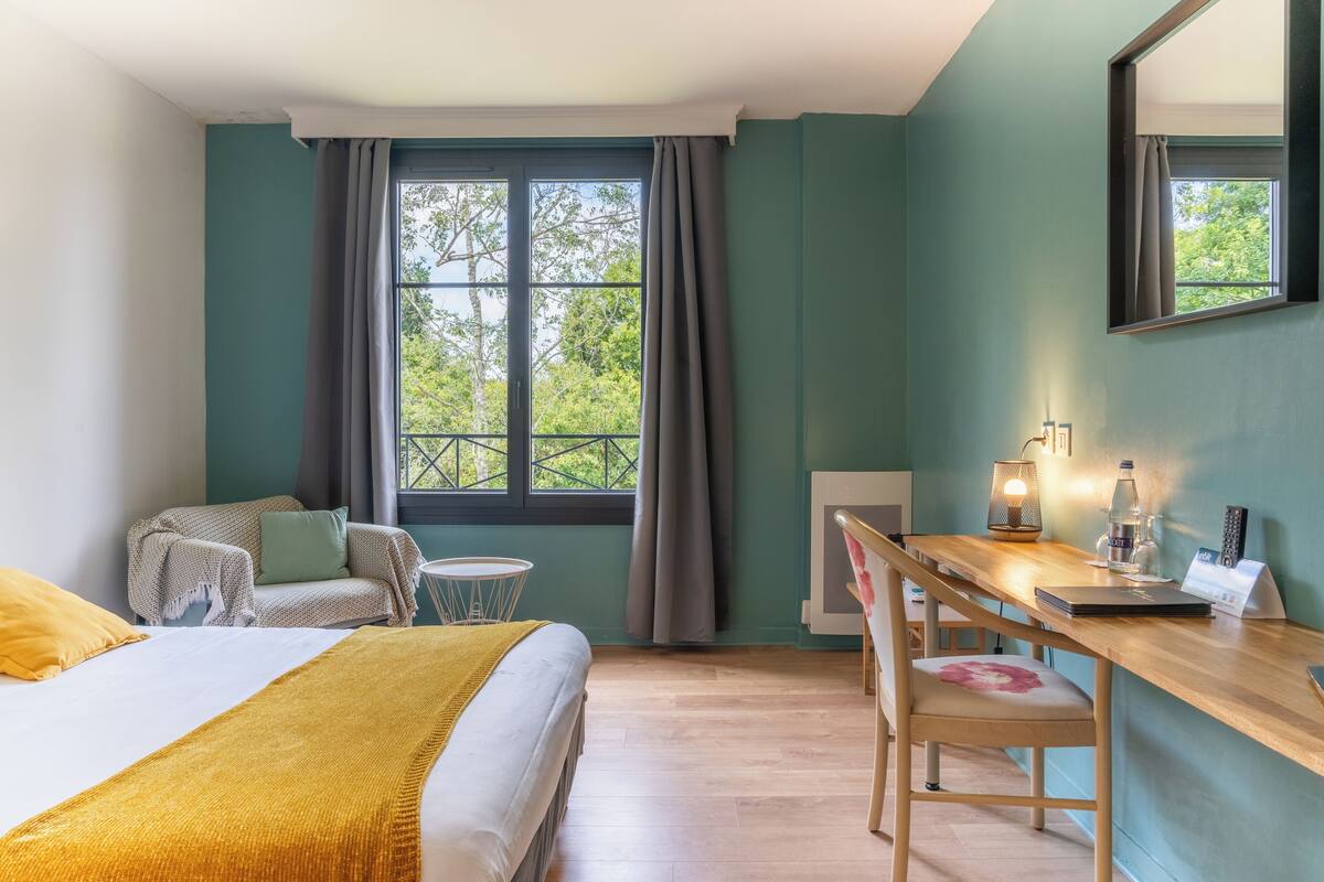 Una habitación con una cama, un escritorio y una silla. Hay una ventana con cortinas, una lámpara y una botella en el escritorio.