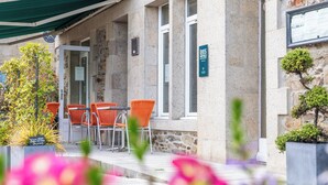 Een hotelentree met stenen muren, een overdekt terras met oranje stoelen, en een bord met de naam van het hotel.