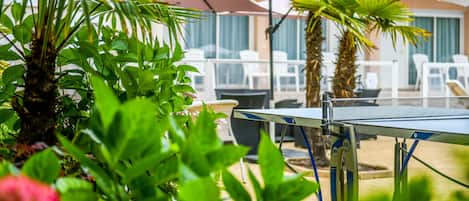 A table tennis table in a garden area with white lounge chairs and umbrellas, surrounded by lush greenery and palm trees.