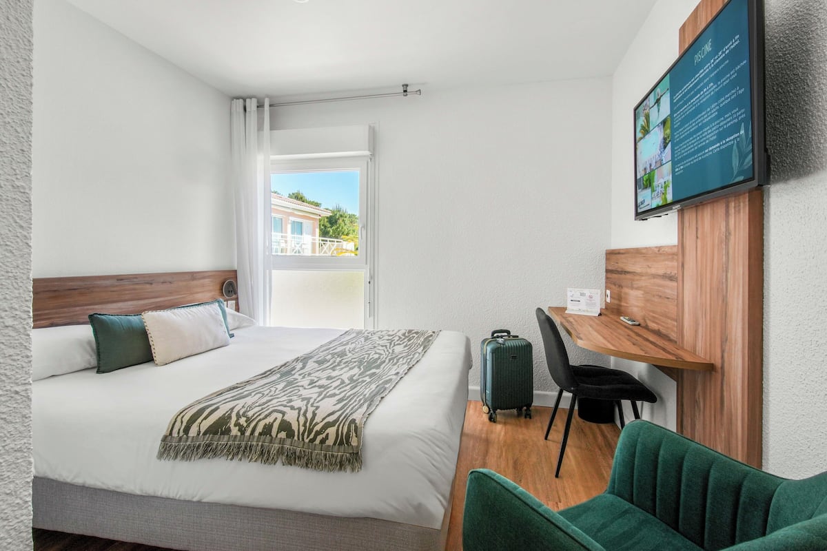 A hotel room with a bed, a desk, a chair, a suitcase, and a TV mounted on the wall.