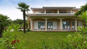 A two-story house with a covered patio area, a well-maintained lawn, and tropical plants. - Hotel La Forestière (Biscarrosse)