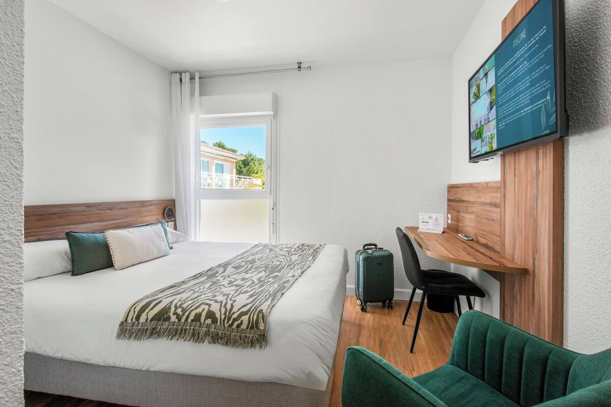 A hotel room with a bed, a desk, a chair, a suitcase, and a TV mounted on the wall.