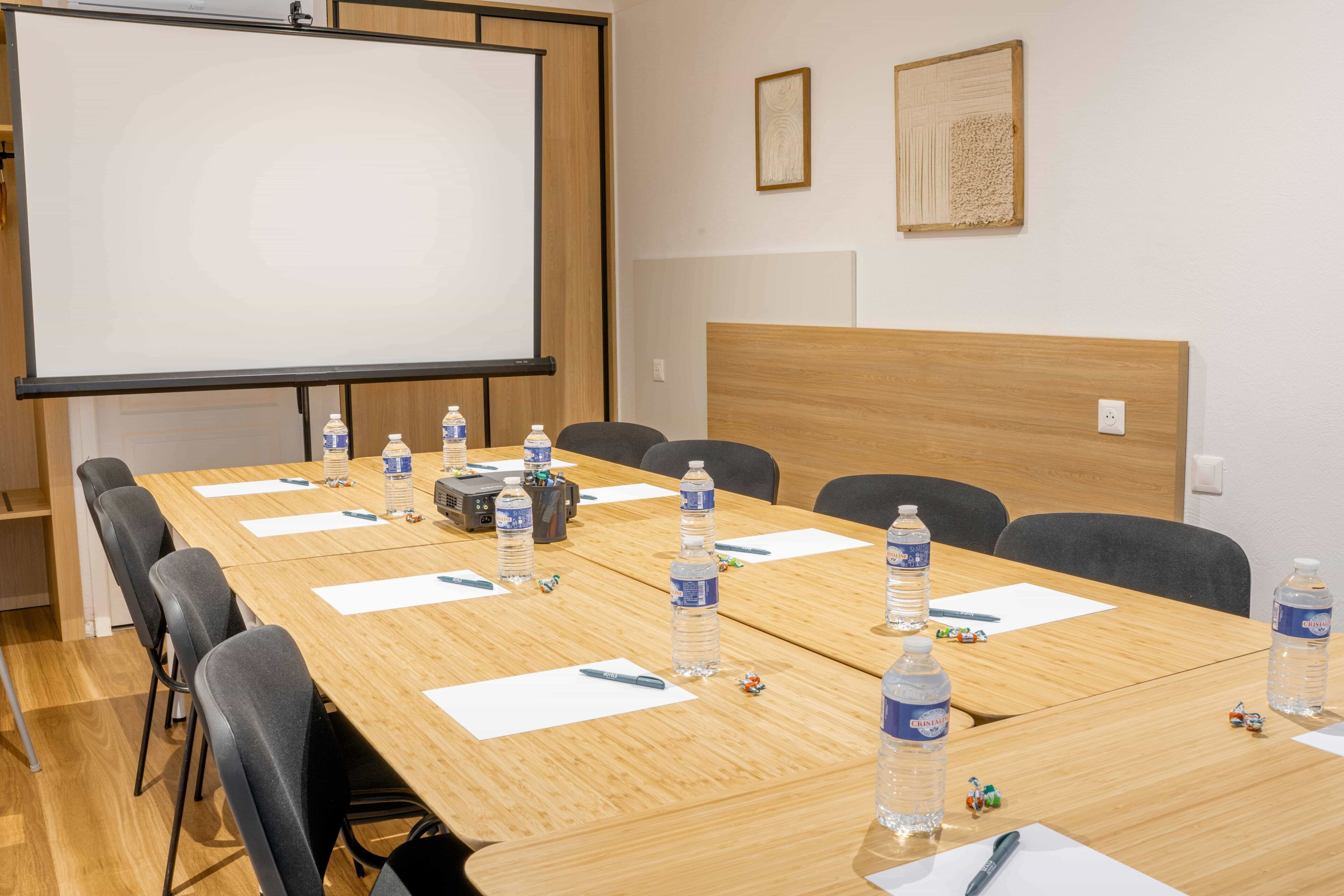 Un salón de conferencias con una larga mesa, sillas, una pantalla de proyector, botellas de agua y una pizarra.