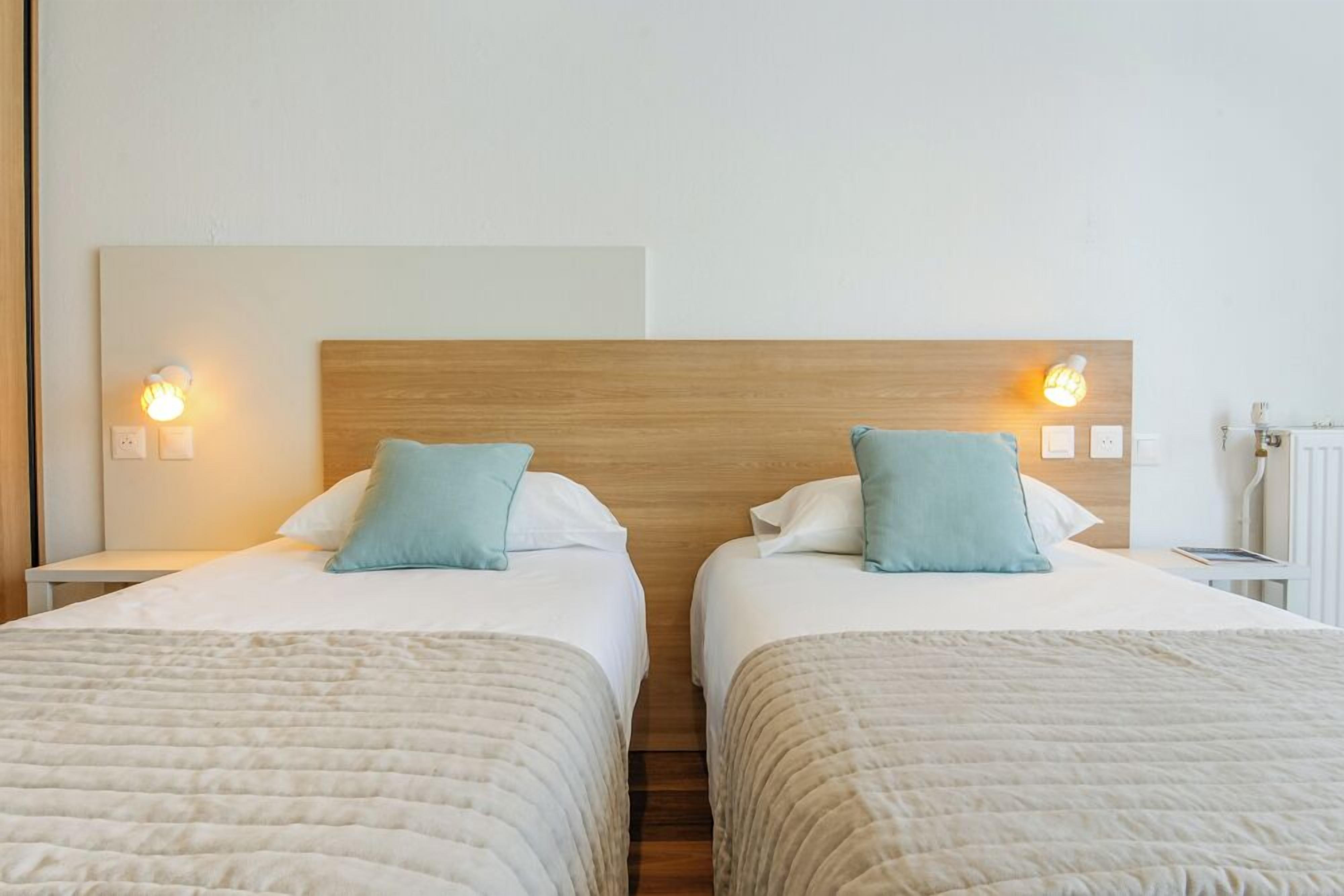 A hotel room with two beds, each with a wooden headboard, white bedding, and a blue pillow. There are bedside lamps and a white radiator.