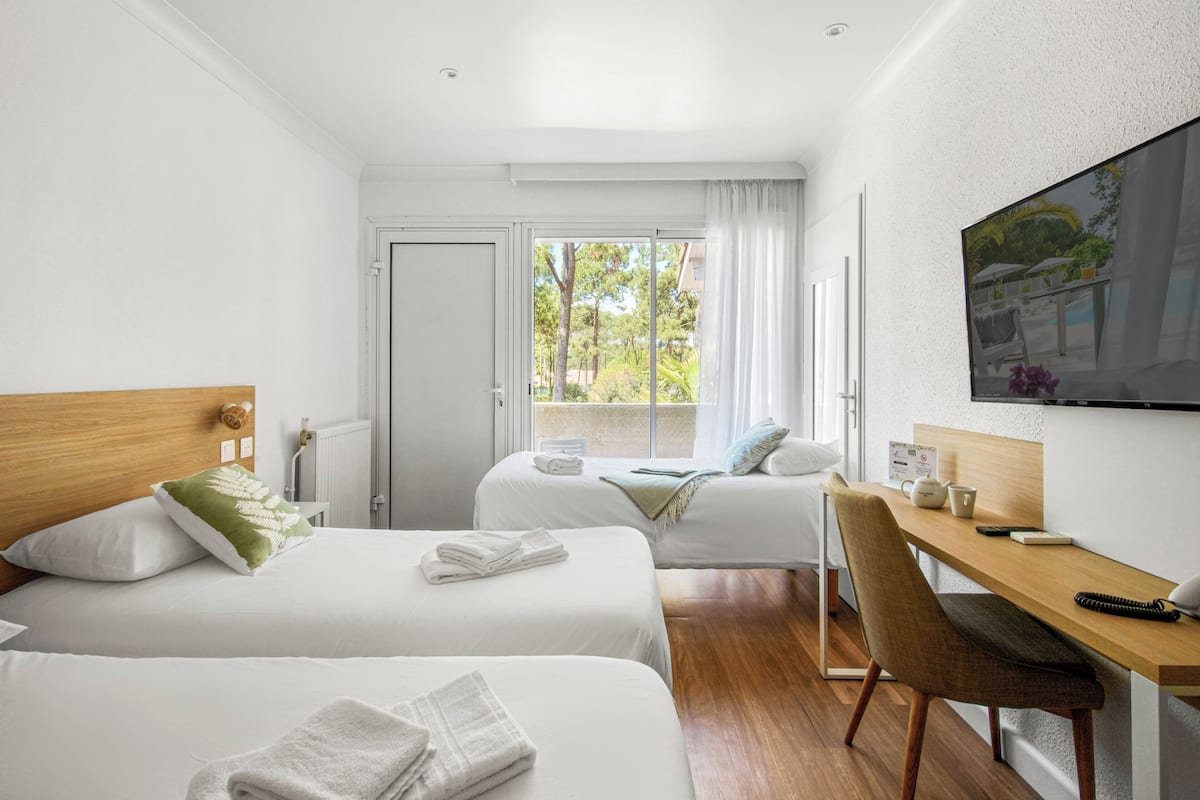 A hotel room with two beds, a desk, a chair, a TV, and a window with a view of greenery.