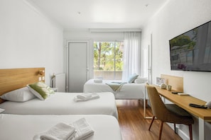 A hotel room with two beds, a desk, a chair, a TV, and a window with a view of greenery.