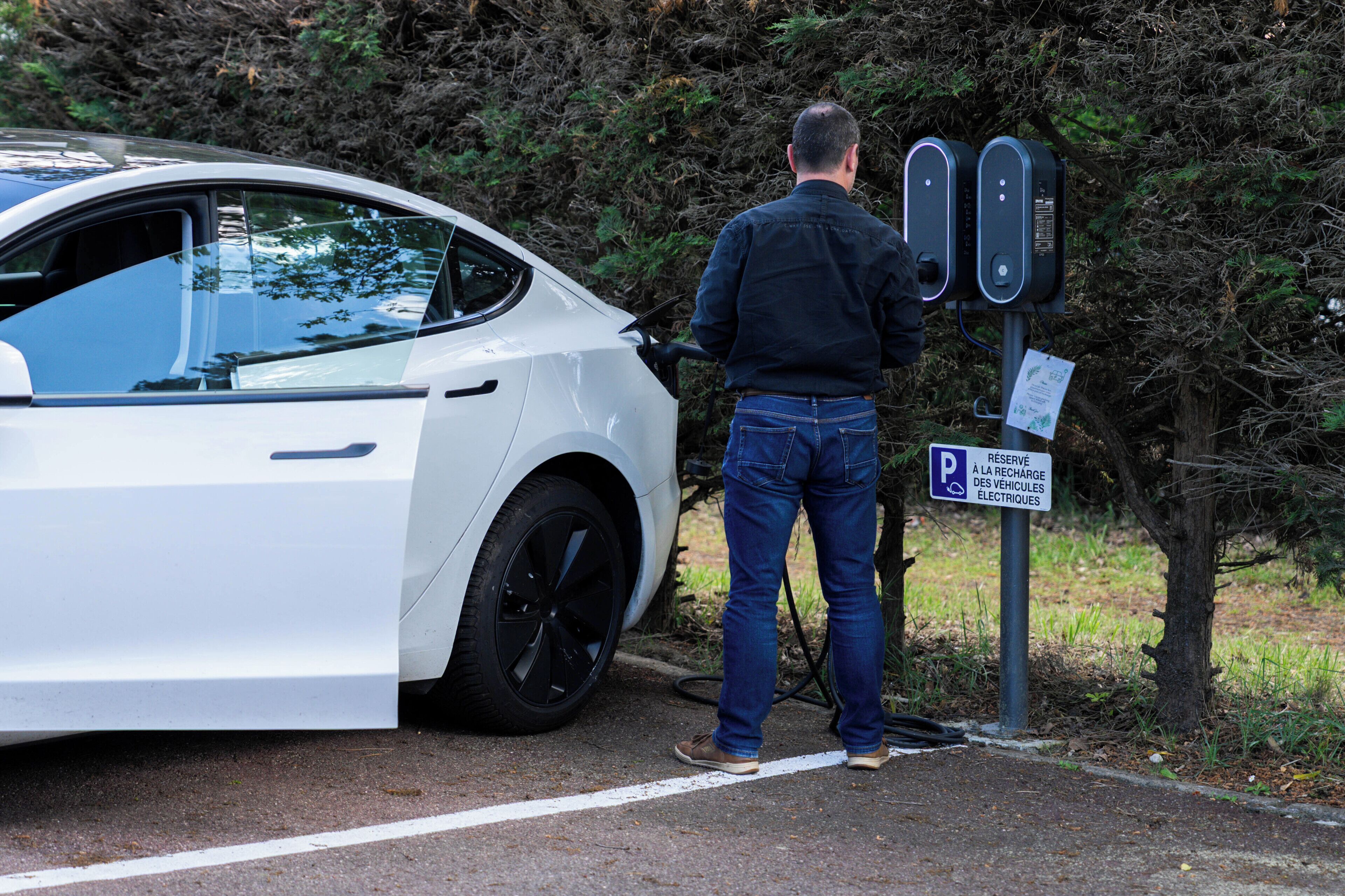 A white car parked at an electric vehicle charging station with a man standing beside it.