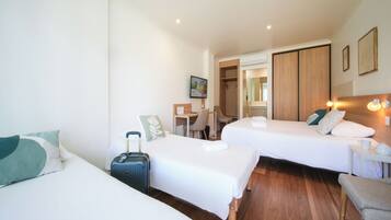 Habitación de hotel con dos camas, un escritorio, una silla, una maleta y una mesita de noche con lámpara.