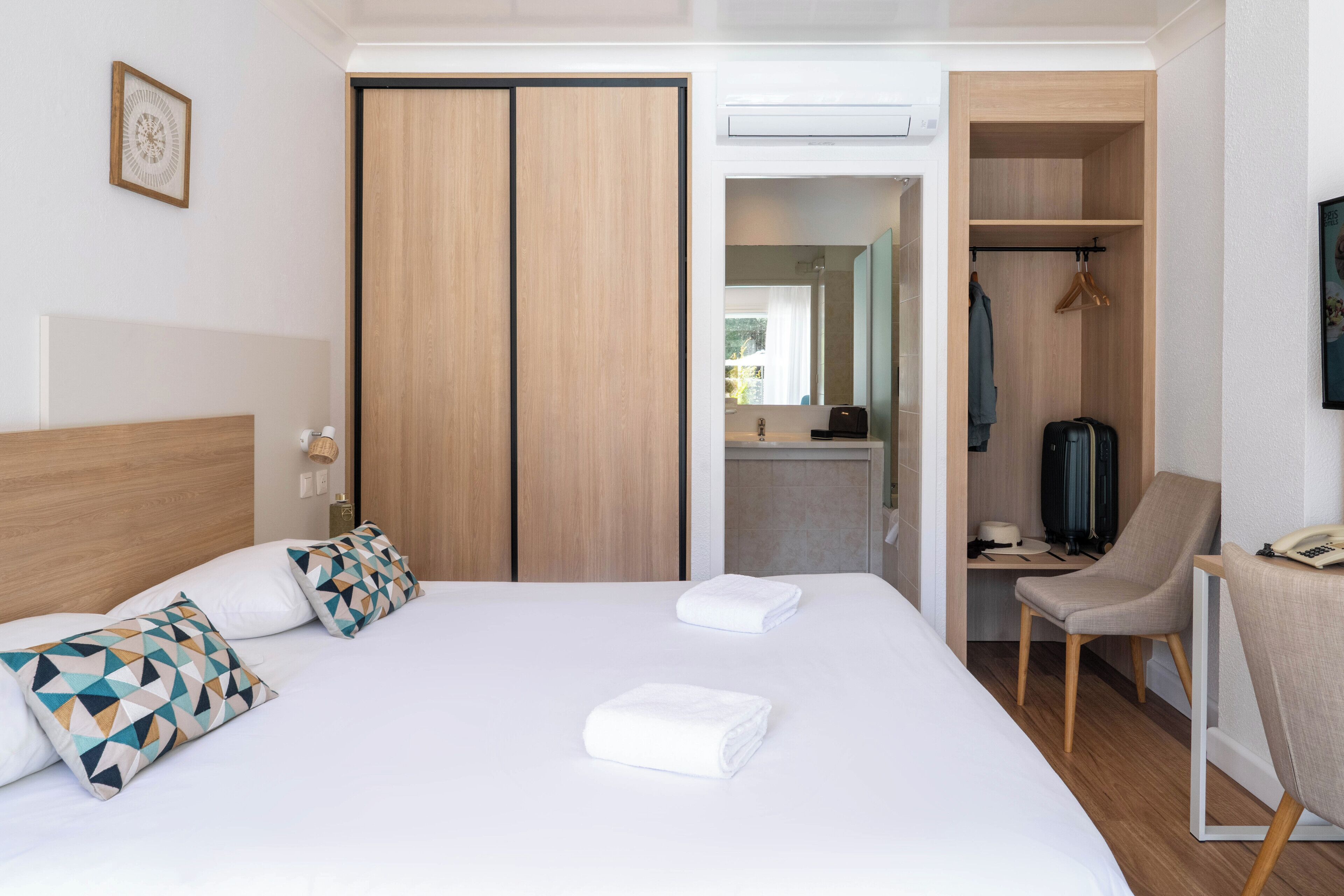A hotel room with a bed, a desk, a chair, a wardrobe, and a bathroom visible through an open door.