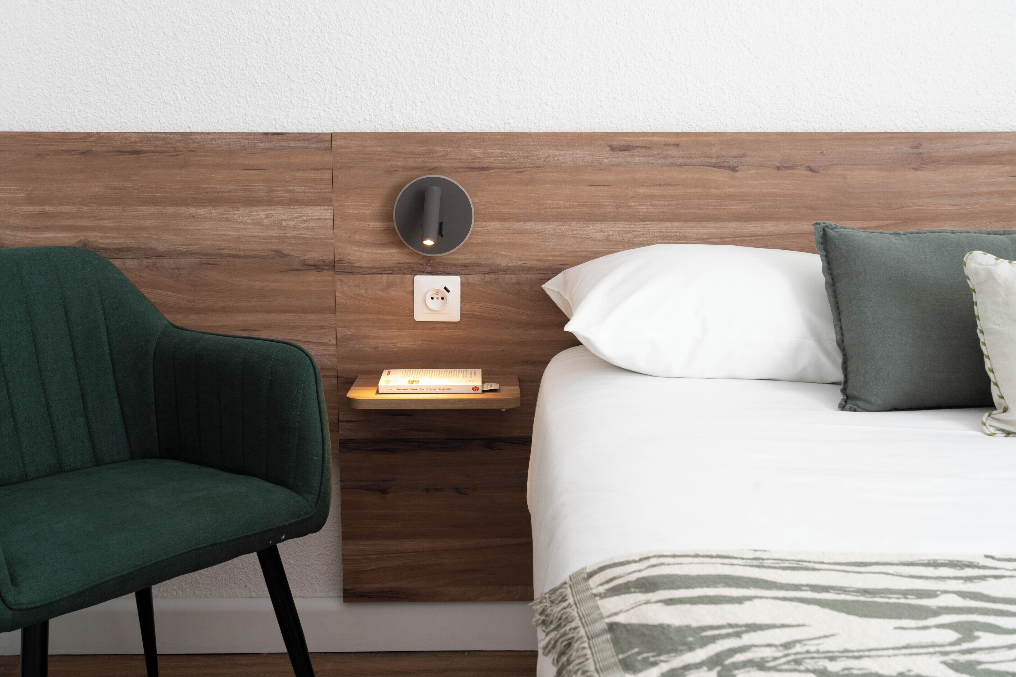 A neatly arranged hotel room with a wooden headboard, a green chair, a bedside table with a book, a white pillow, and a patterned throw.