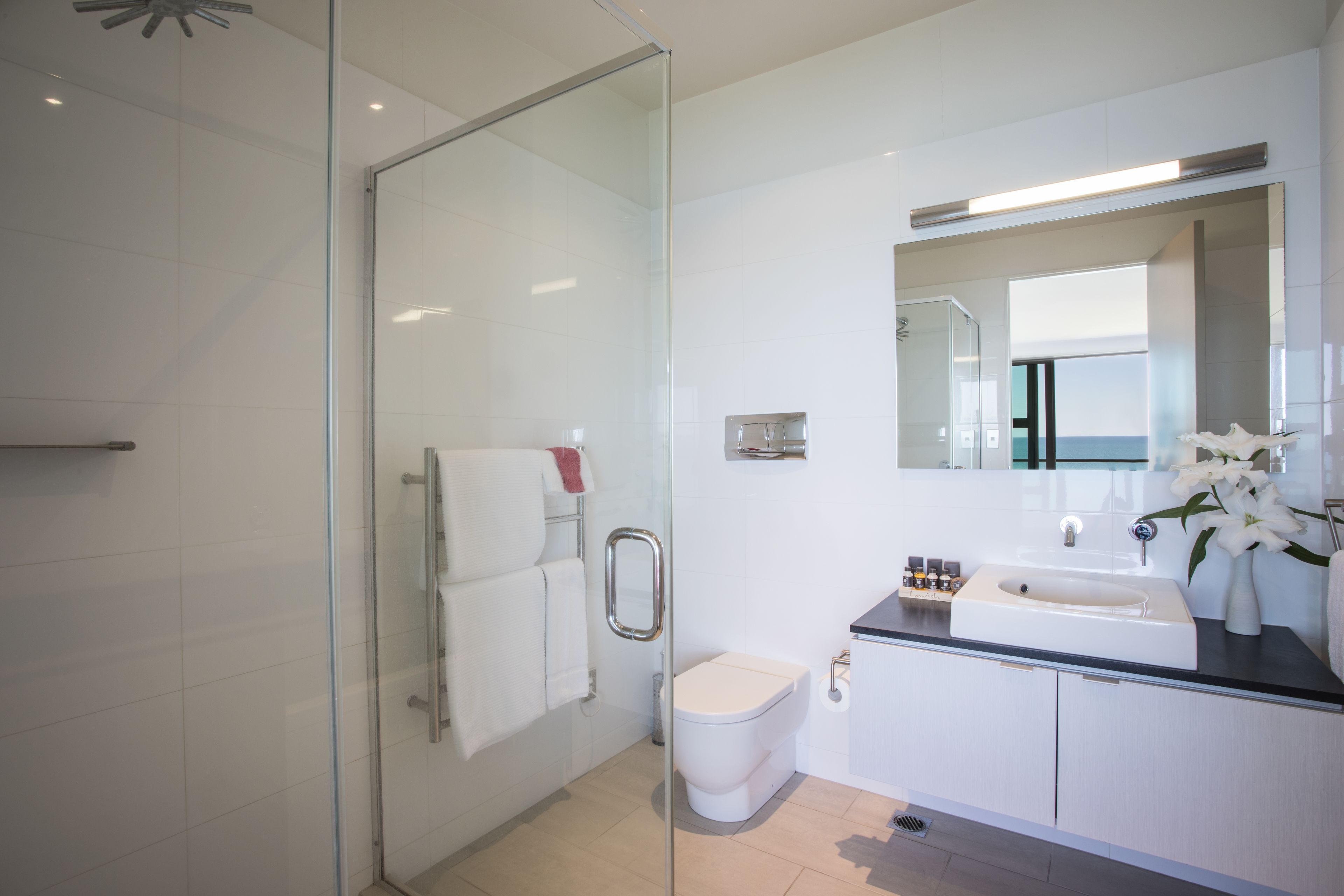 Un baño moderno con cabina de ducha de vidrio, inodoro blanco, lavabo con espejo y un jarrón con flores.