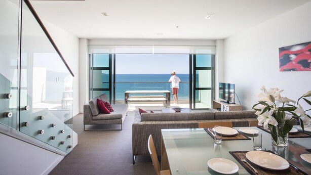 A modern living room with a glass wall overlooking the sea, a dining area with a table set for four, and a sofa with red cushions. - Doubtless Bay Villas (Cable Bay)