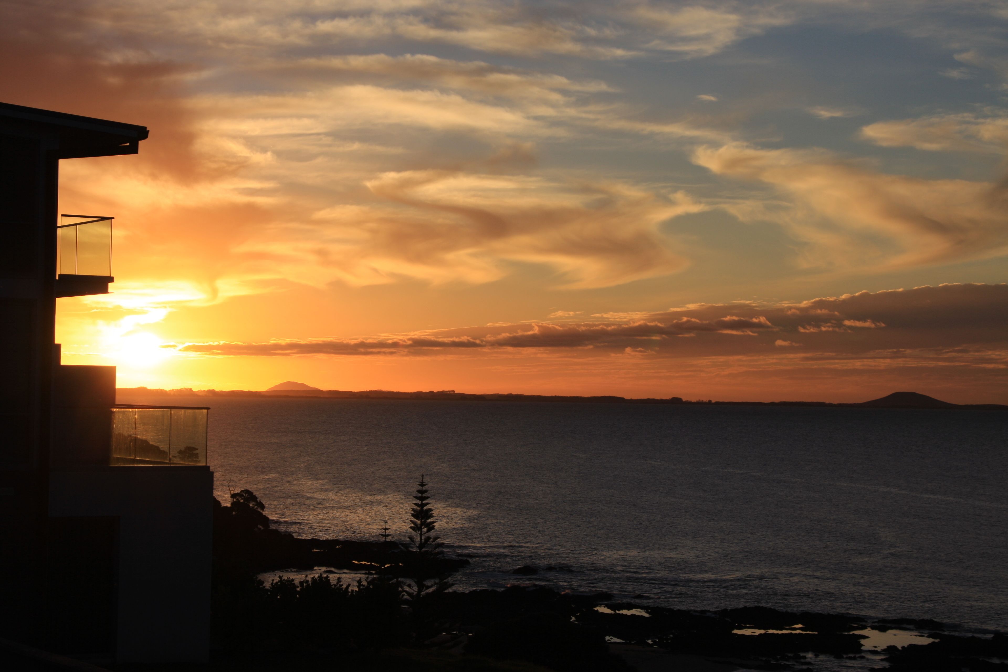 Vista del atardecer desde un balcón con vista al mar, y una montaña distante en el horizonte.