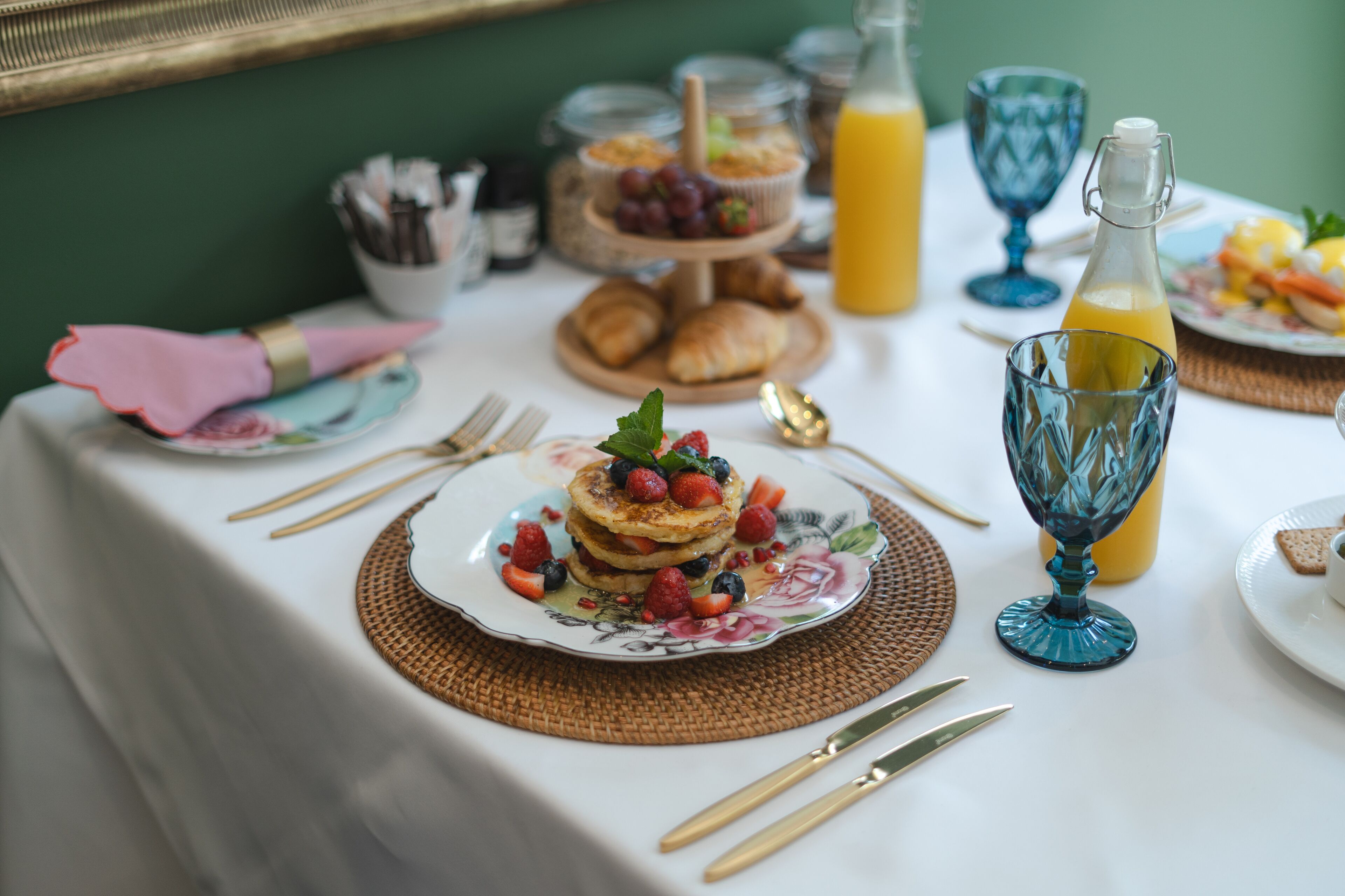 Una mesa de desayuno con panqueques, frutas, cruasanes y una botella de jugo de naranja.