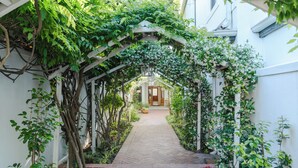 Un estrecho sendero de ladrillo que conduce a una puerta de madera, rodeado de abundante vegetación y plantas con flores blancas.