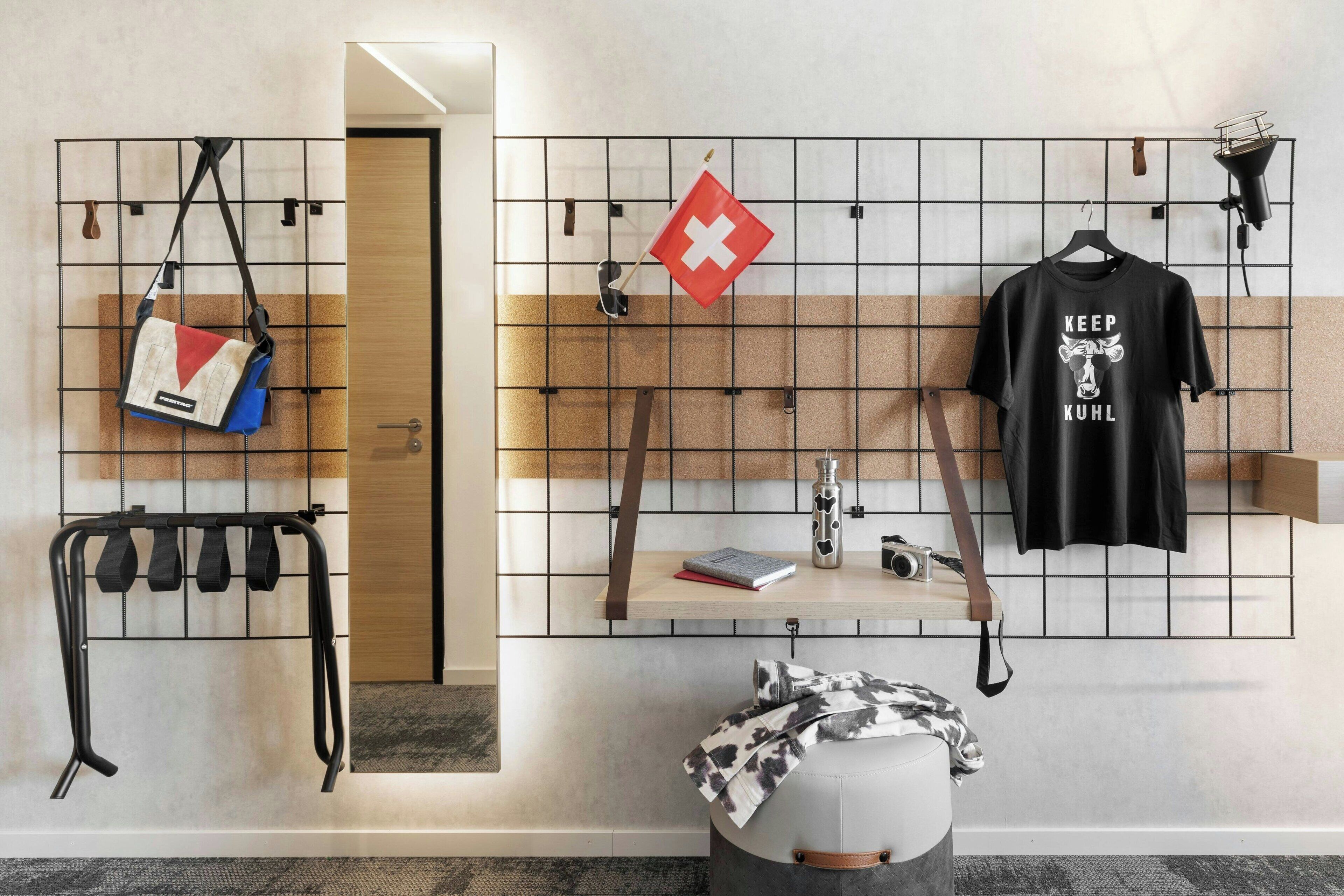 Une chambre d’hôtel avec un mur en forme de grille, un porte-bagages suspendu, un drapeau suisse, un t-shirt noir avec un slogan, un appareil photo et une petite table avec un livre et une bouteille.