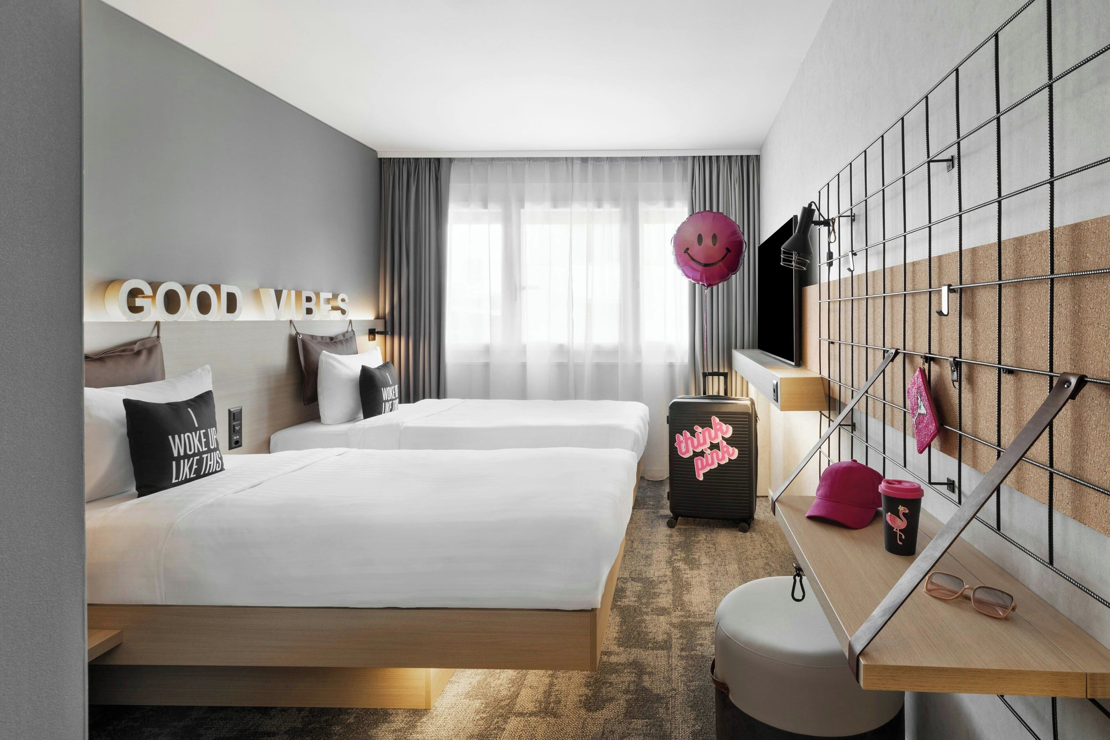 A modern hotel room with two beds, a desk, a suitcase, and a wall-mounted TV.