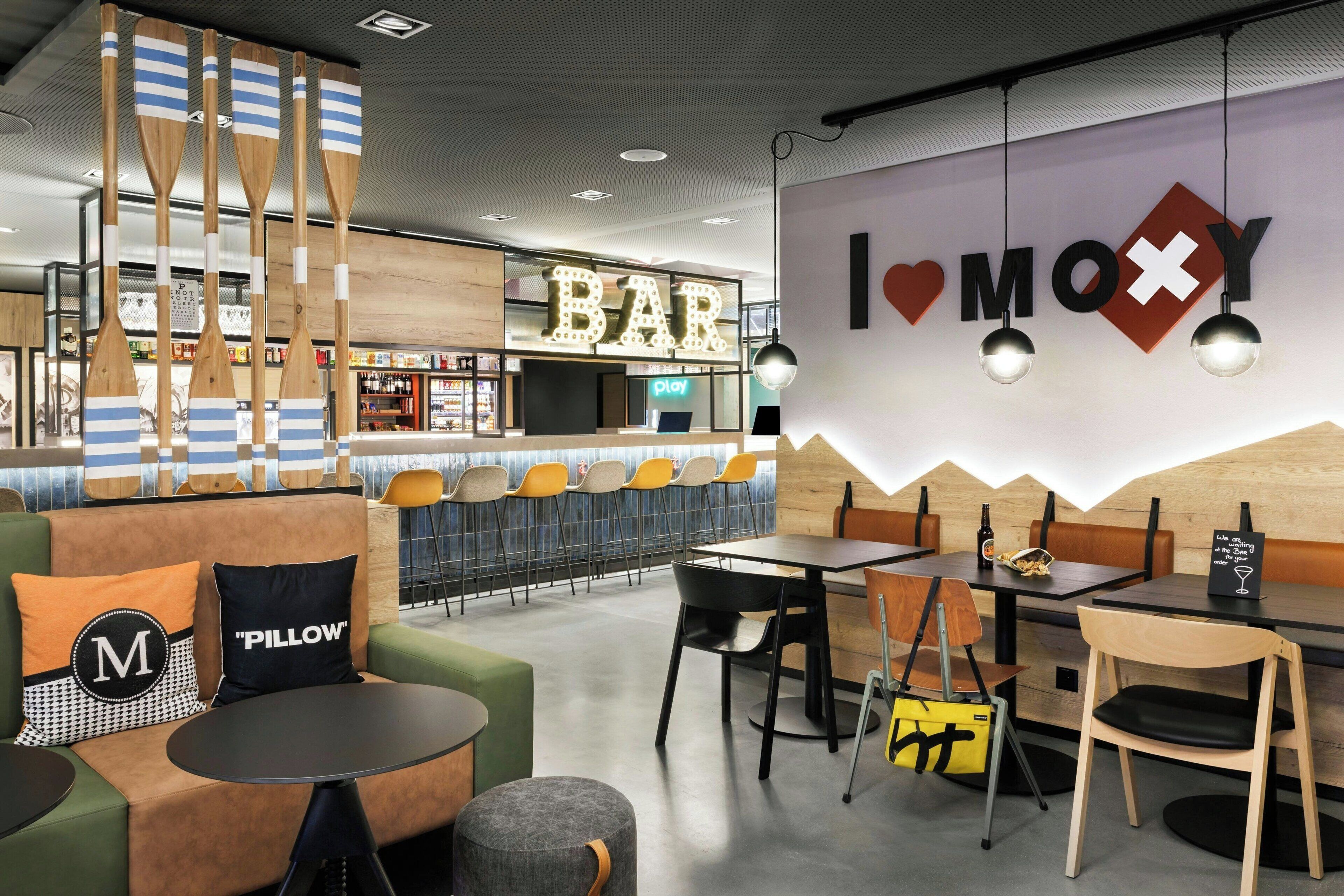 A modern bar area with a 'BAR' sign, seating arrangements, and a 'MOXY' sign on the wall.