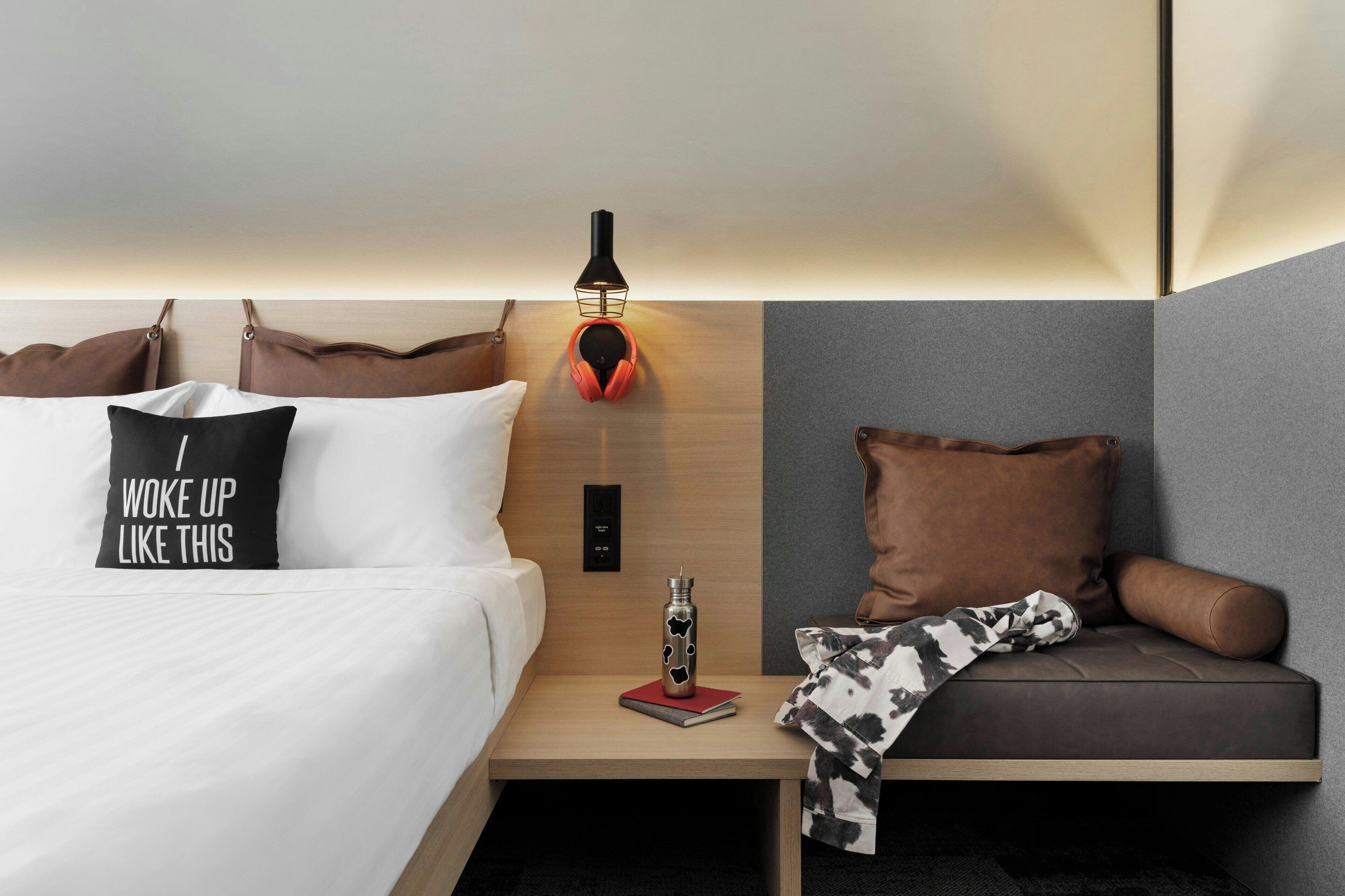 Ein modernes Hotelzimmer mit einem ordentlich bezogenen Bett, einem Nachttisch mit Lampe, einem roten Buch und einer Wasserflasche.