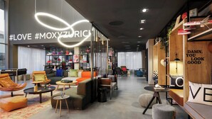 A modern lounge area with sofas, chairs, and a coffee table. There are shelves with books and decorative items. - Moxy Zurich (Zürich)