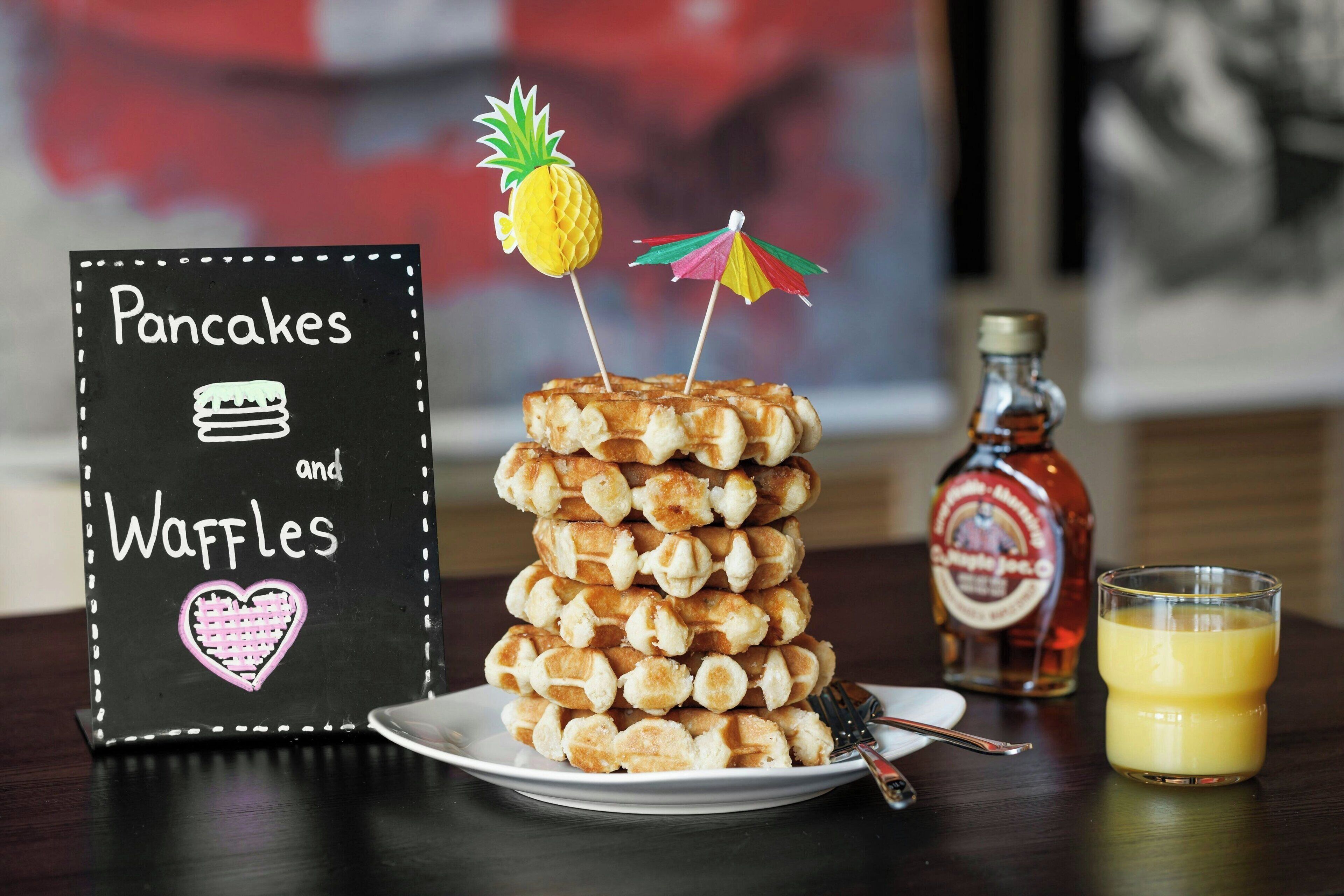A stack of pancakes and waffles with a sign indicating the menu, a bottle of syrup, and a glass of orange juice on a table.