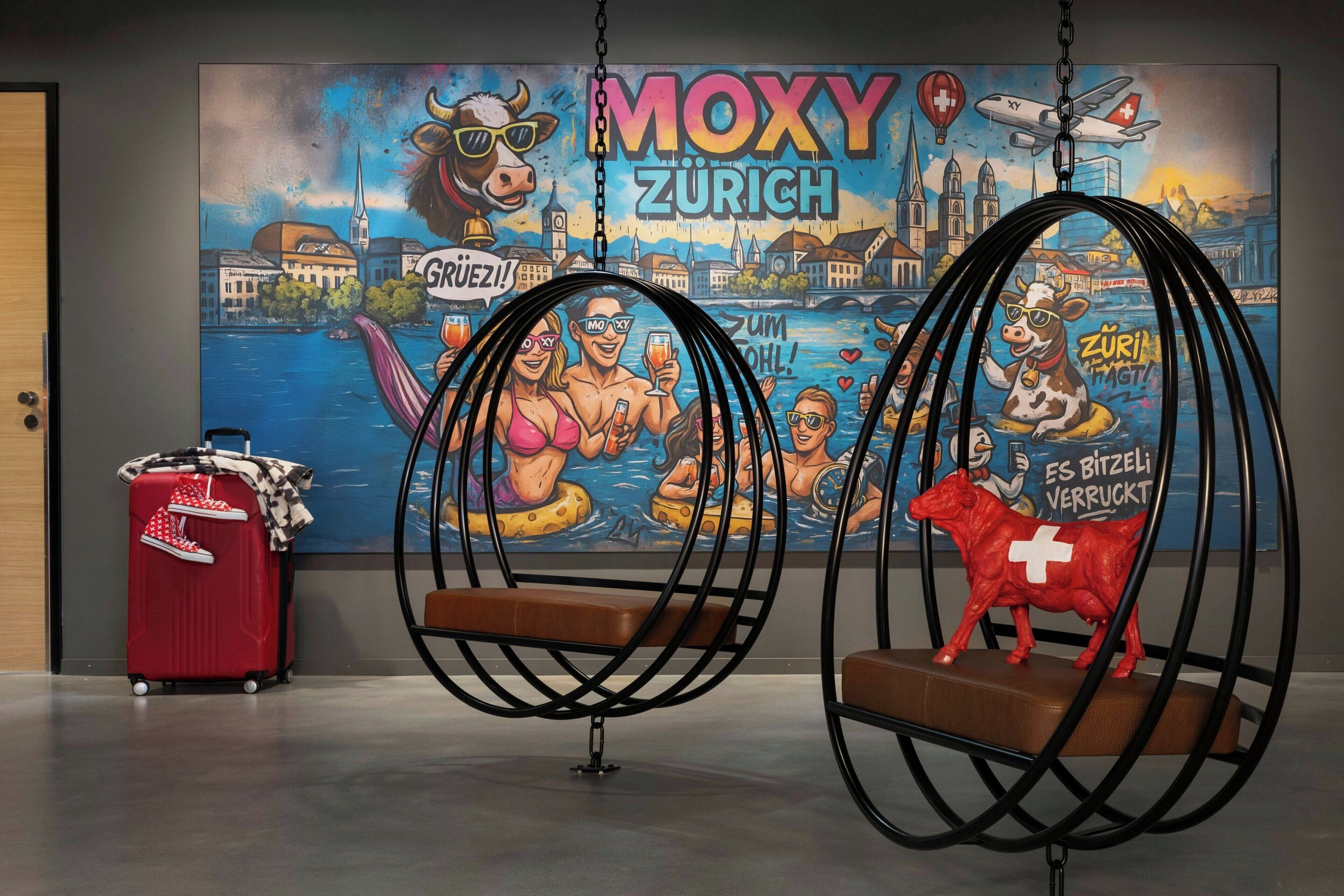 A room with two black swing chairs, a red suitcase, and a mural of MoXY Zürich.