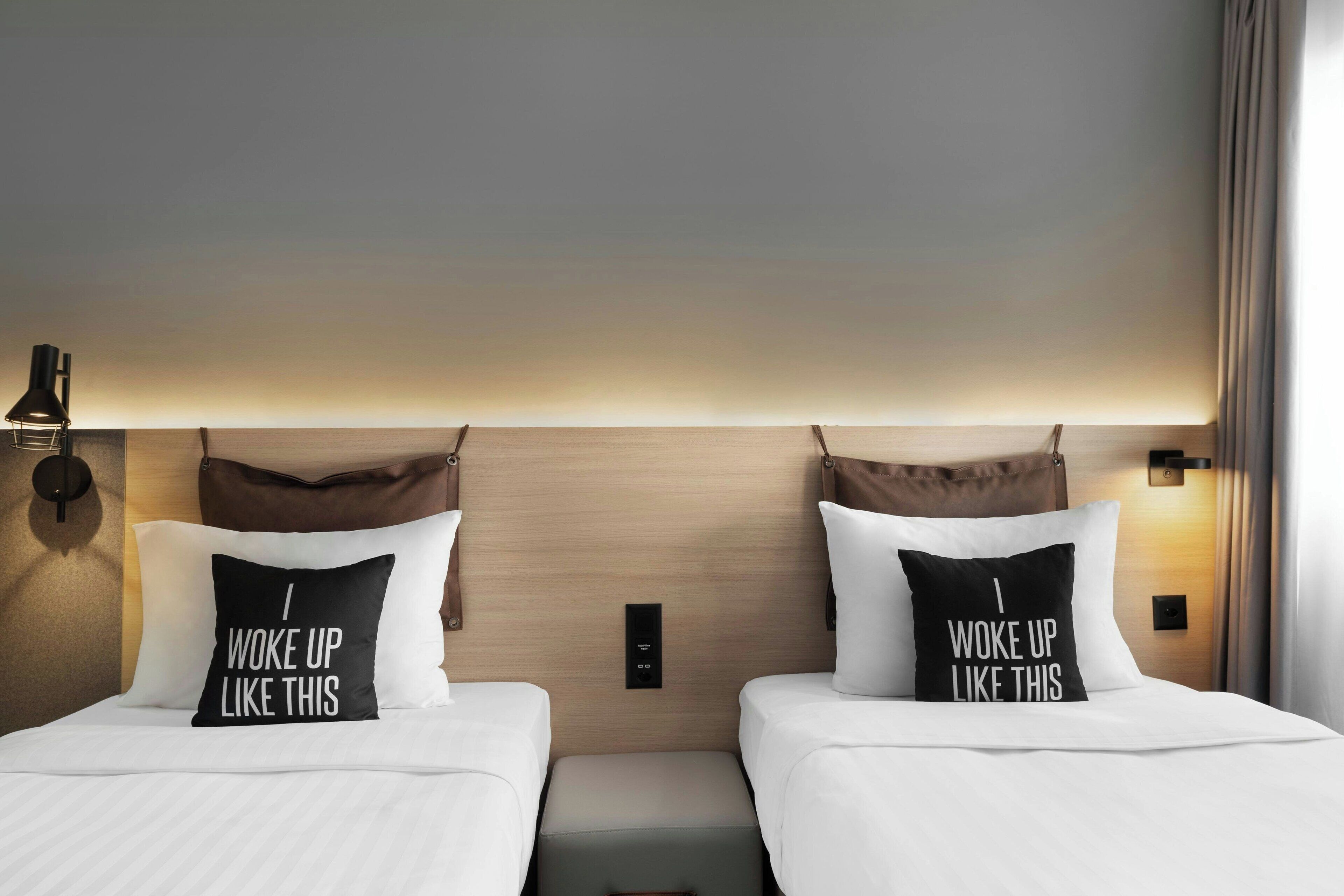 Habitación de hotel con dos camas, cada una con un almohadón que dice 'Me levanté así'.