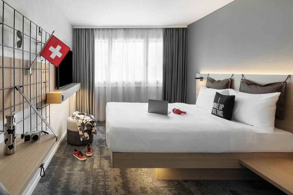 A modern hotel room with a bed, a laptop, a Swiss flag, a TV, and a window with curtains.