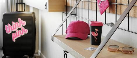 A wooden shelf with a pink cap, a flamingo mug, and a pair of glasses. A black suitcase with "think pink" text is visible in the background.