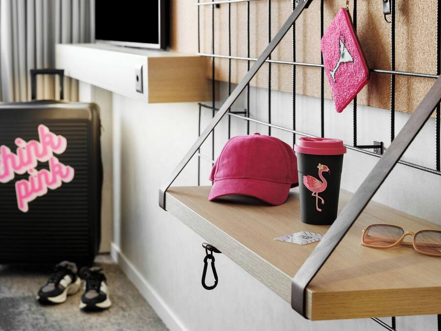 Ein Holzregal mit einer rosafarbenen Kappe, einer Flamingo-Tasse und einer Brille. Im Hintergrund ist ein schwarzer Koffer mit der Aufschrift "think pink" zu sehen.