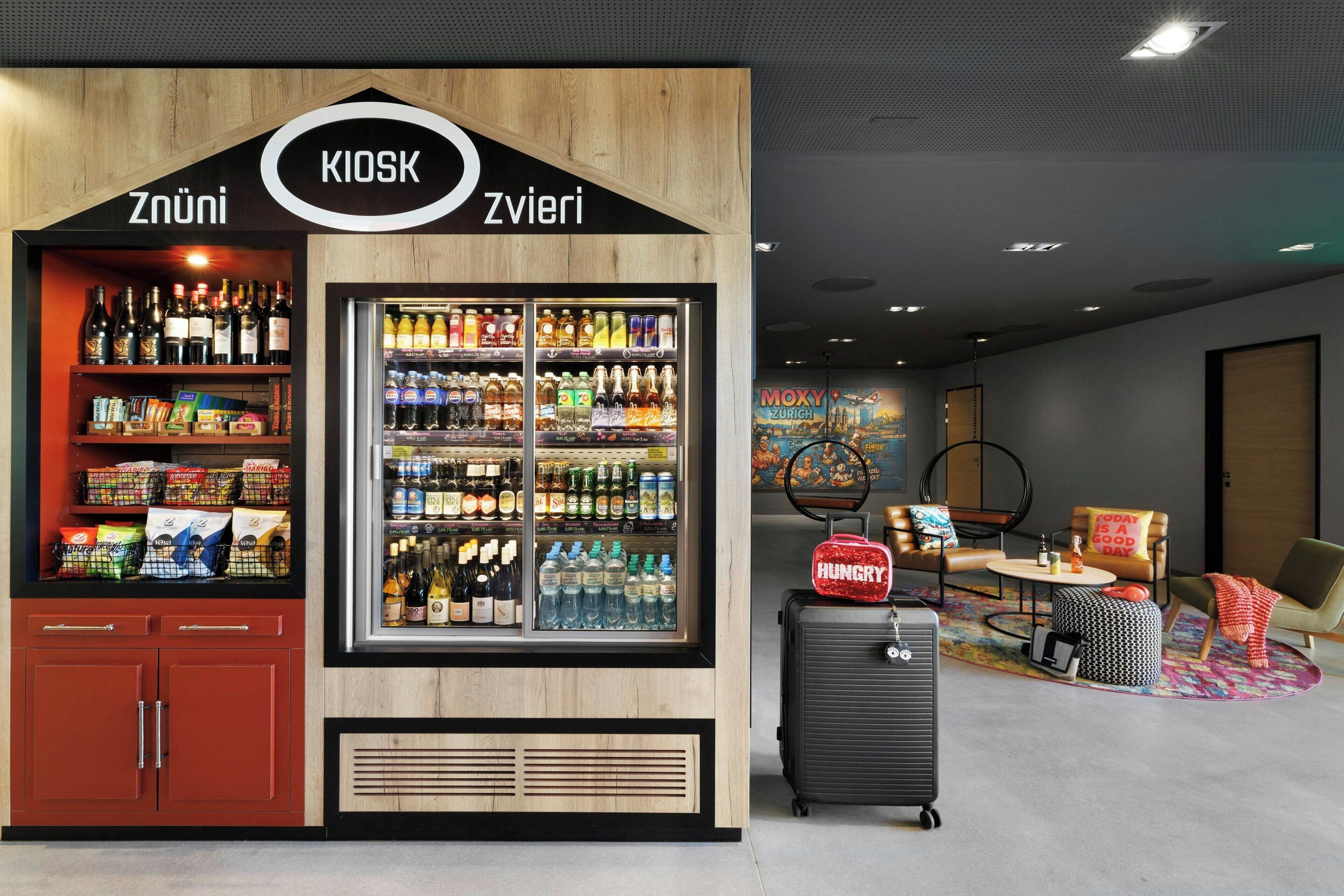 A kiosk with shelves of snacks and beverages, a luggage trolley, and a seating area with a colorful rug.