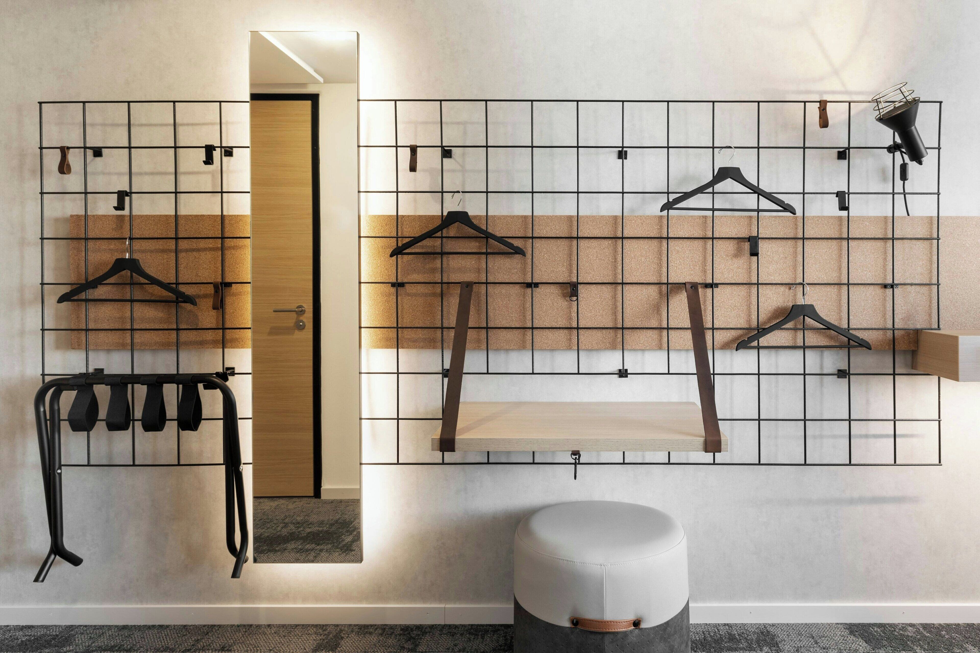 Une pièce avec un panneau mural en forme de grille sur lequel sont fixés plusieurs cintres noirs, une porte en bois et un petit pouf rond.