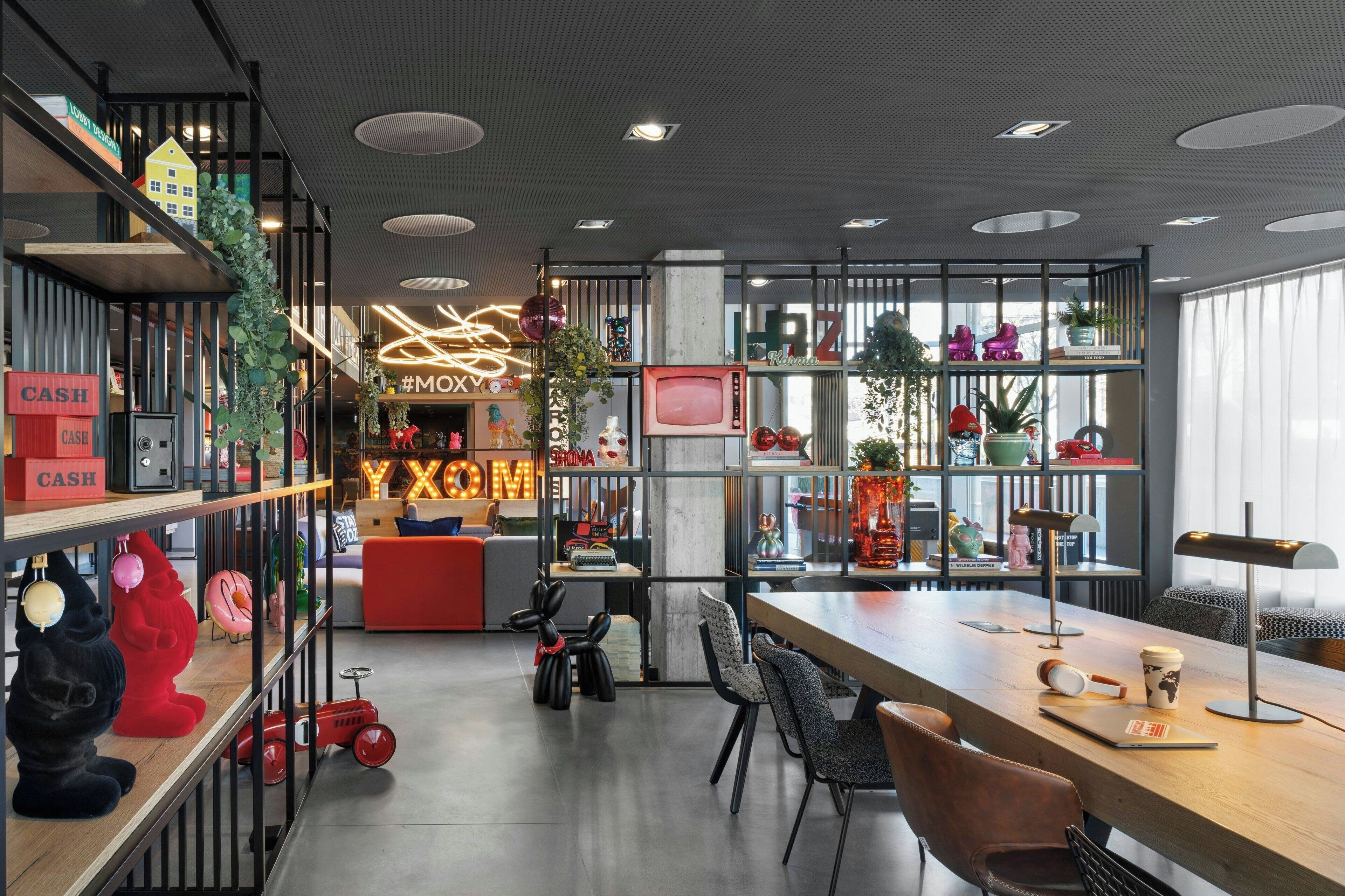 A modern interior with a long wooden table, black chairs, and a red sofa. There are shelves with various items and a neon sign.