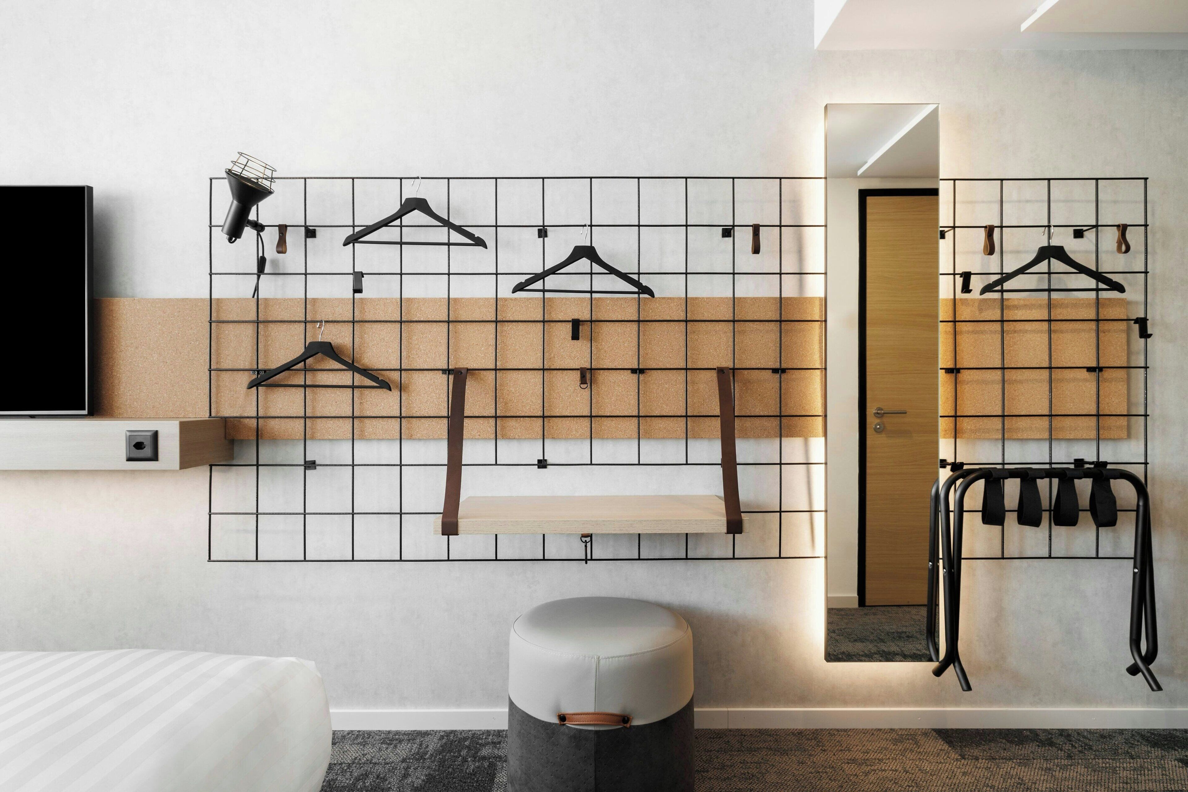 Una habitación de hotel moderna con una cama, un televisor montado, un perchero sujeto a la pared, un taburete pequeño y un espejo.