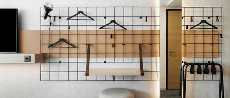 A modern hotel room with a bed, a mounted TV, a wall-mounted rack with hangers, a small stool, and a mirror.
