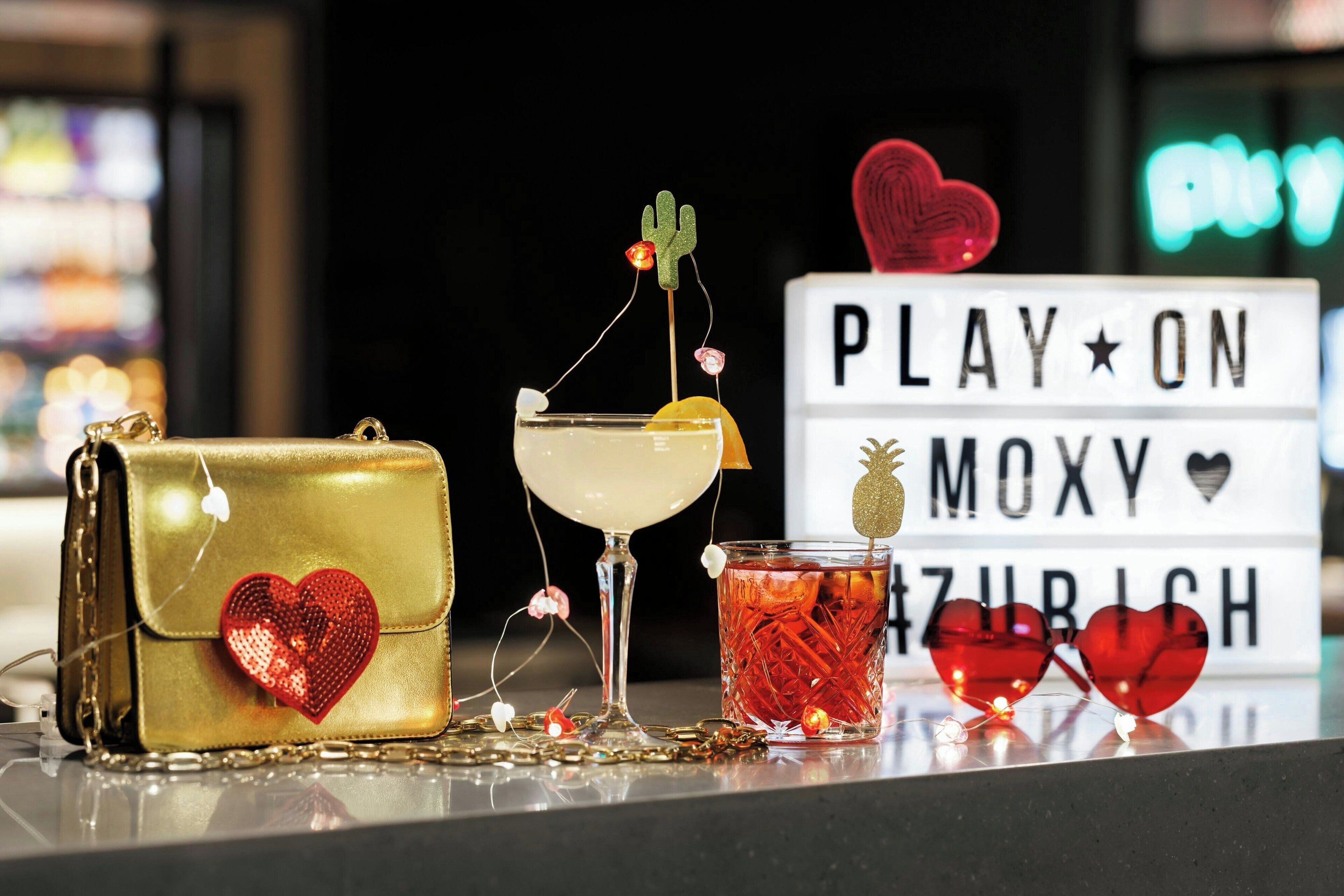 A table with a gold handbag, a cocktail glass, a drink with a cactus decoration, and a sign that reads "PLAY ON MOXY HEARTZURICH".