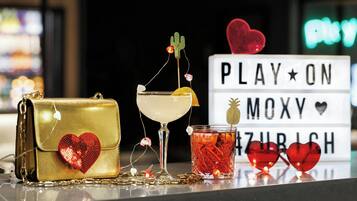 A table with a gold handbag, a cocktail glass, a drink with a cactus decoration, and a sign that reads "PLAY ON MOXY HEARTZURICH".