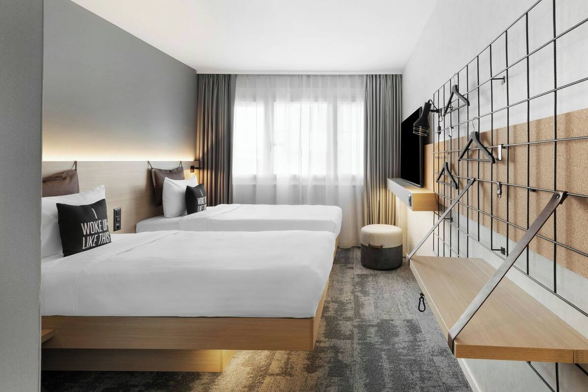 A modern hotel room with two beds, a seating area, a TV mounted on a wall, and a staircase.